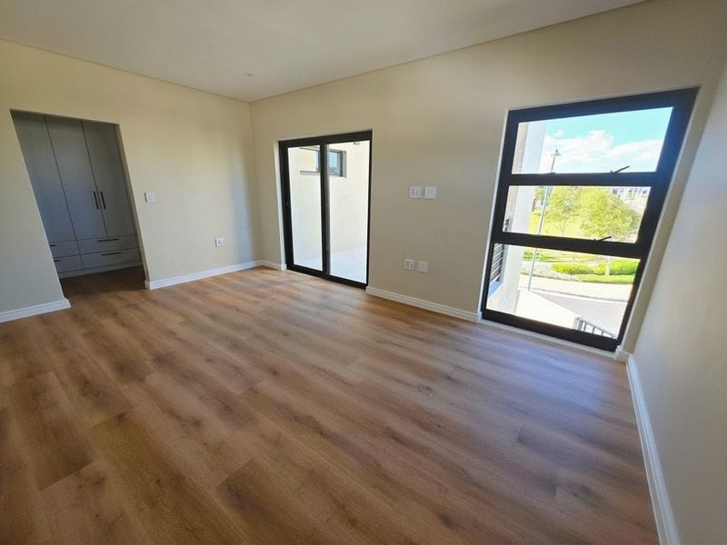 A bright, modern bedroom featuring laminate flooring, built-in cupboards, and large windows providing natural light and outdoor views.
