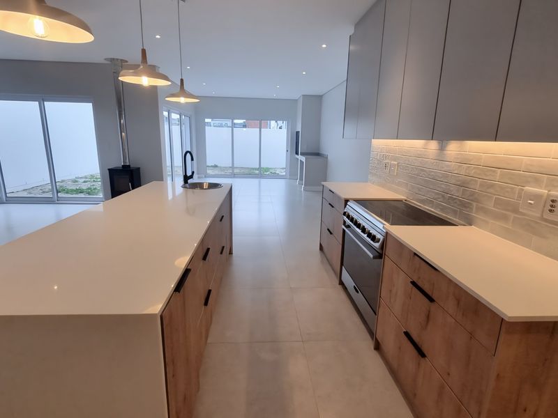 Modern open-plan kitchen featuring a large island, sleek cabinetry, integrated appliances, and a wood-burning fireplace, flowing into a bright living area with sliding doors.