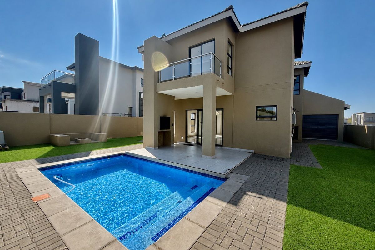 Rear view of the property featuring a swimming pool, covered patio with built-in braai, and an upper-level balcony with glass balustrades.