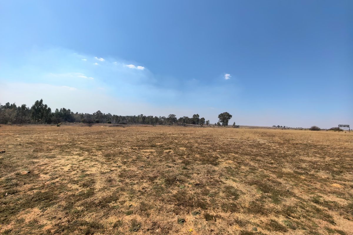 A wide-angle view of a vacant residential land plot featuring dry grass and distant trees under a clear blue sky.