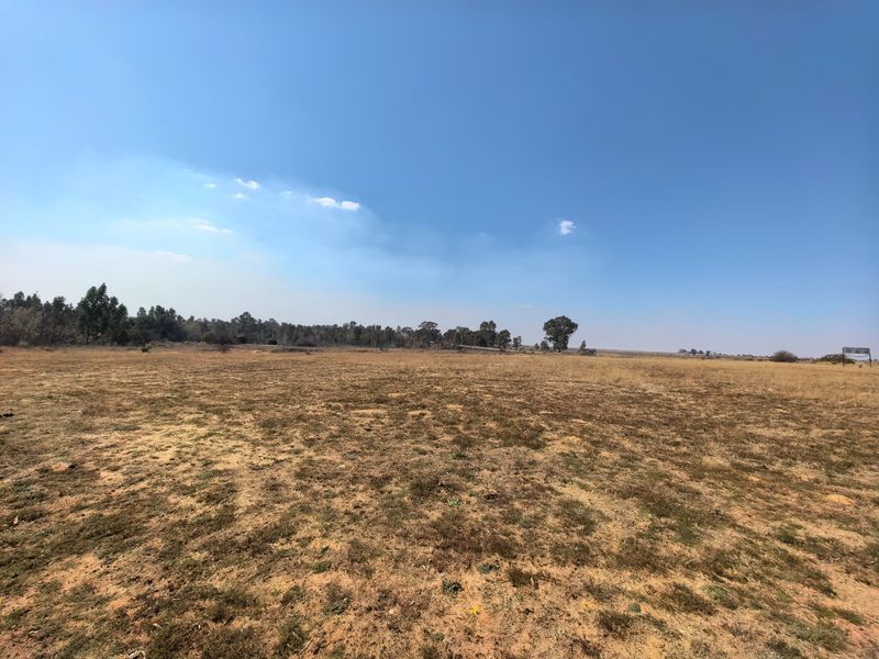 A wide-angle view of a vacant residential land plot featuring dry grass and distant trees under a clear blue sky.
