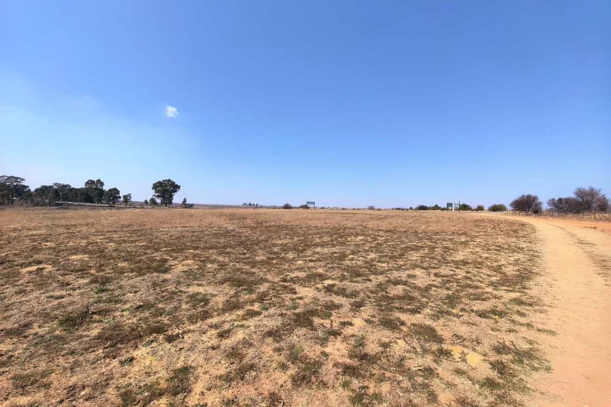 A wide-angle view of a vacant residential plot featuring dry grass, a dirt access road, and perimeter fencing under a clear blue sky.