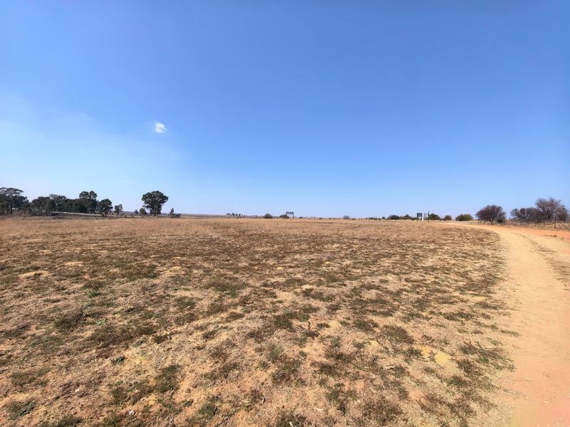 A wide-angle view of a vacant residential plot featuring dry grass, a dirt access road, and perimeter fencing under a clear blue sky.