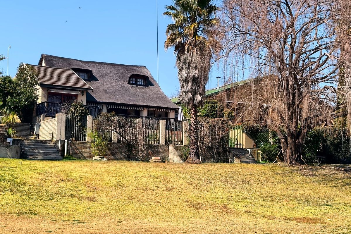 Rear view of a house featuring a traditional thatched roof, a raised balcony, and a brick retaining wall overlooking a large lawn area.