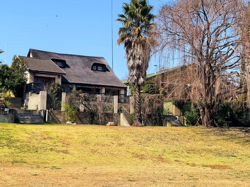 Rear view of a house featuring a traditional thatched roof, a raised balcony, and a brick retaining wall overlooking a large lawn area.