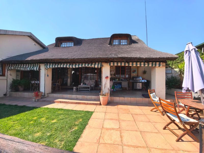 Rear view of a traditional thatched-roof house featuring a spacious tiled patio, retractable awnings, and dormer windows overlooking a garden area.