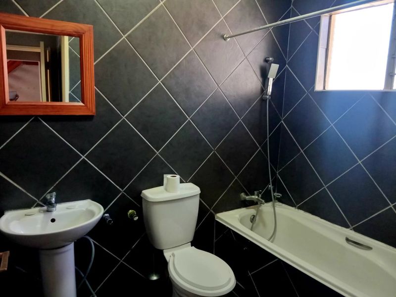 A bathroom featuring dark diamond-patterned wall tiles, a pedestal basin, a toilet, and a bathtub with a handheld shower attachment.
