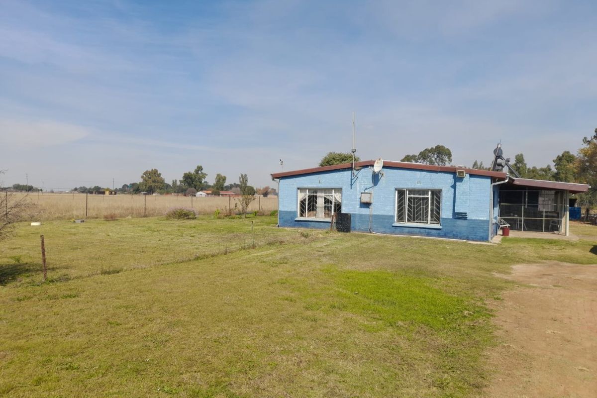 Rear view of a single-story farm dwelling featuring a blue painted brick exterior, metal security bars on windows, and an attached covered patio area.