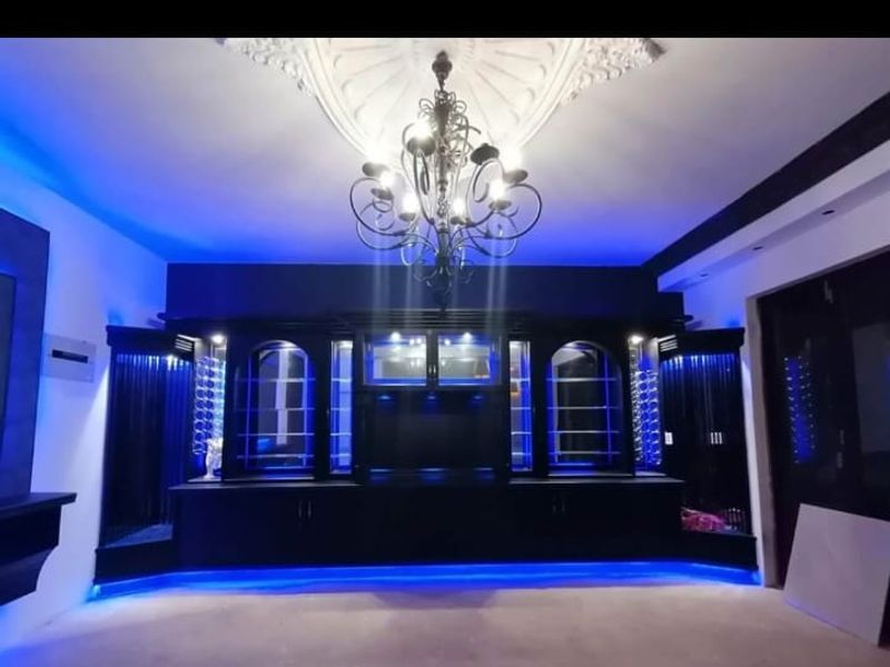 A spacious living area featuring custom built-in cabinetry with integrated blue LED lighting, a decorative ceiling rose, and a classic wrought iron chandelier.
