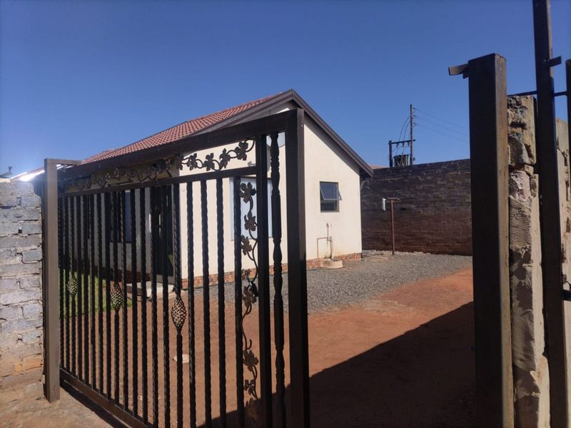 Front view of the property featuring a secure wrought iron gate, gravel driveway, and a tiled roof structure with a brick boundary wall.