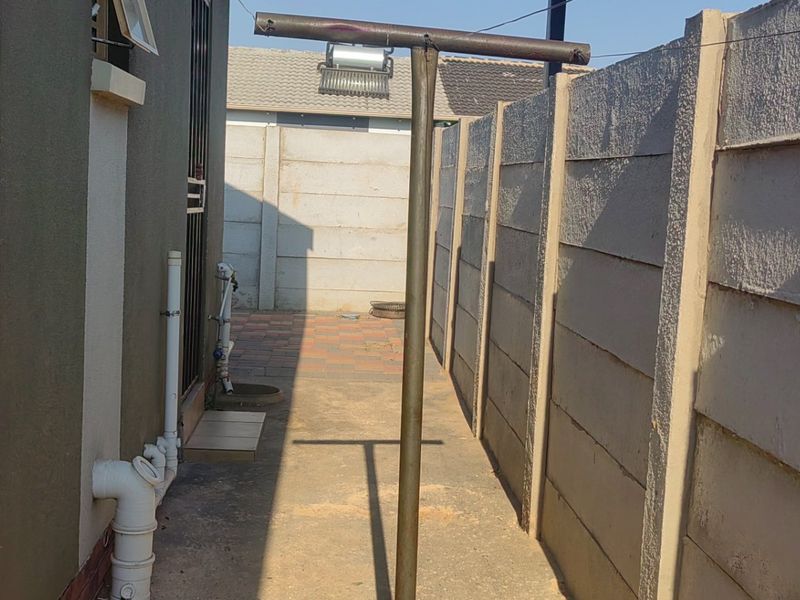 A narrow rear exterior side passage featuring a paved walkway, precast concrete boundary wall, and external plumbing infrastructure.