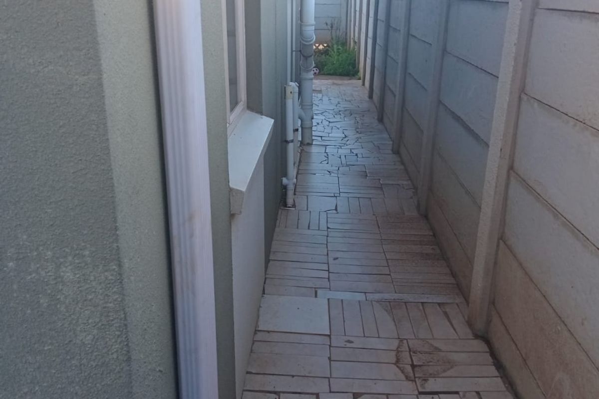 A narrow side walkway featuring paved flooring, enclosed by a precast concrete boundary wall and external building drainage pipes.