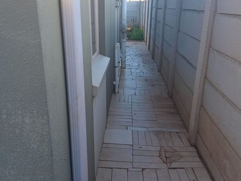 A narrow side walkway featuring paved flooring, enclosed by a precast concrete boundary wall and external building drainage pipes.
