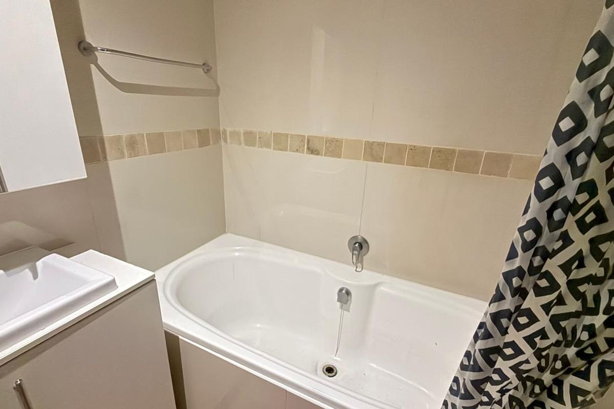 A functional bathroom featuring a built-in bathtub, vanity cabinet with basin, and neutral wall tiling with a decorative border.