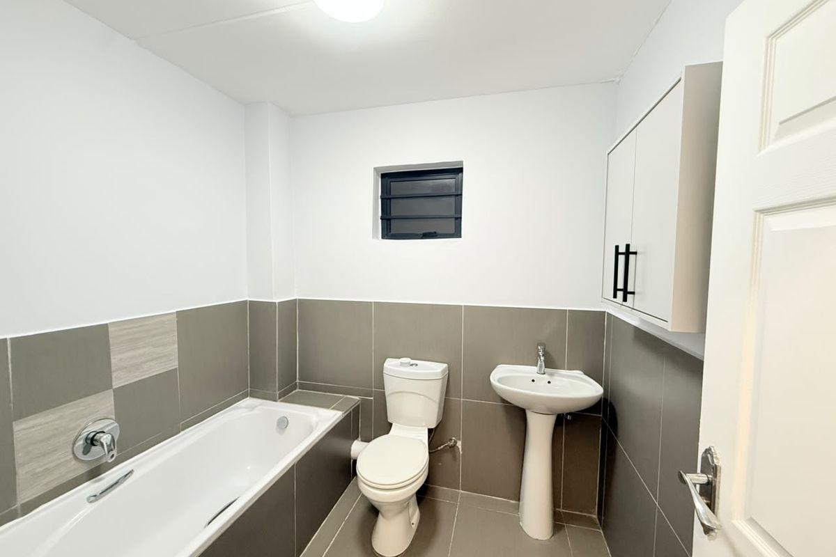 A clean, modern bathroom featuring a bathtub, pedestal sink, toilet, and wall-mounted storage cabinet with neutral grey wall and floor tiling.