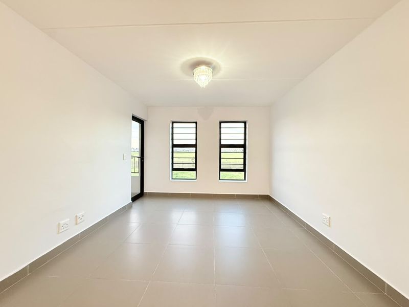 A bright, empty living area featuring neutral tiled flooring, modern aluminium-framed windows, and a glass door leading to an outdoor space.