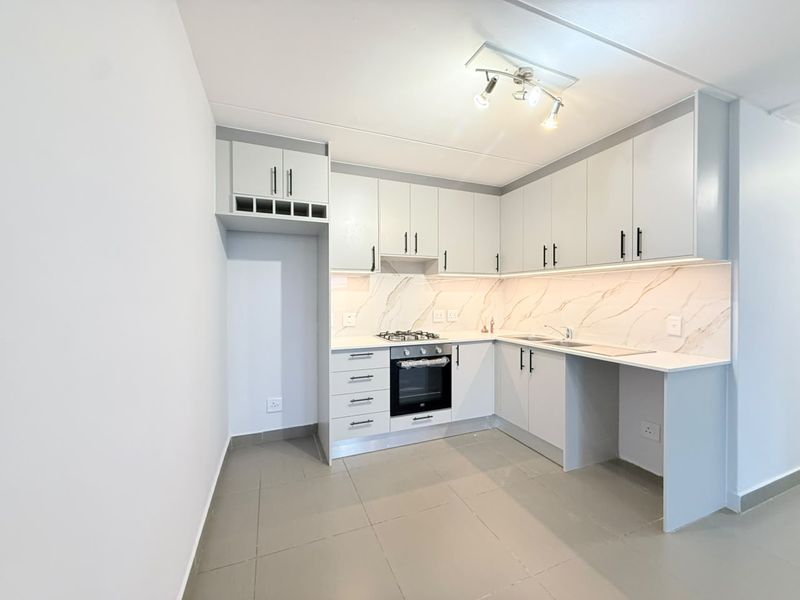 A modern, clean kitchen featuring light grey cabinetry, stone-look backsplash, built-in oven and gas hob, and neutral tiled flooring.
