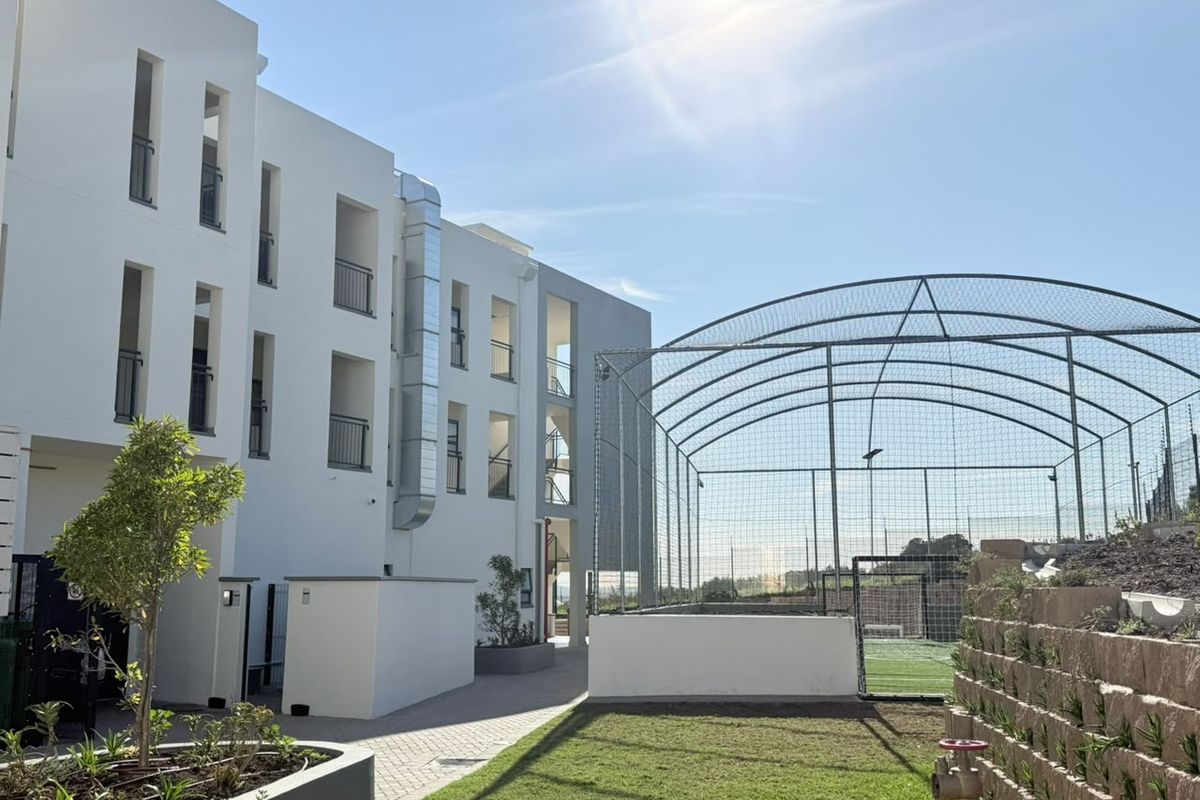 Exterior view of a modern apartment building featuring a paved courtyard, landscaped retaining wall, and an adjacent fenced sports facility.