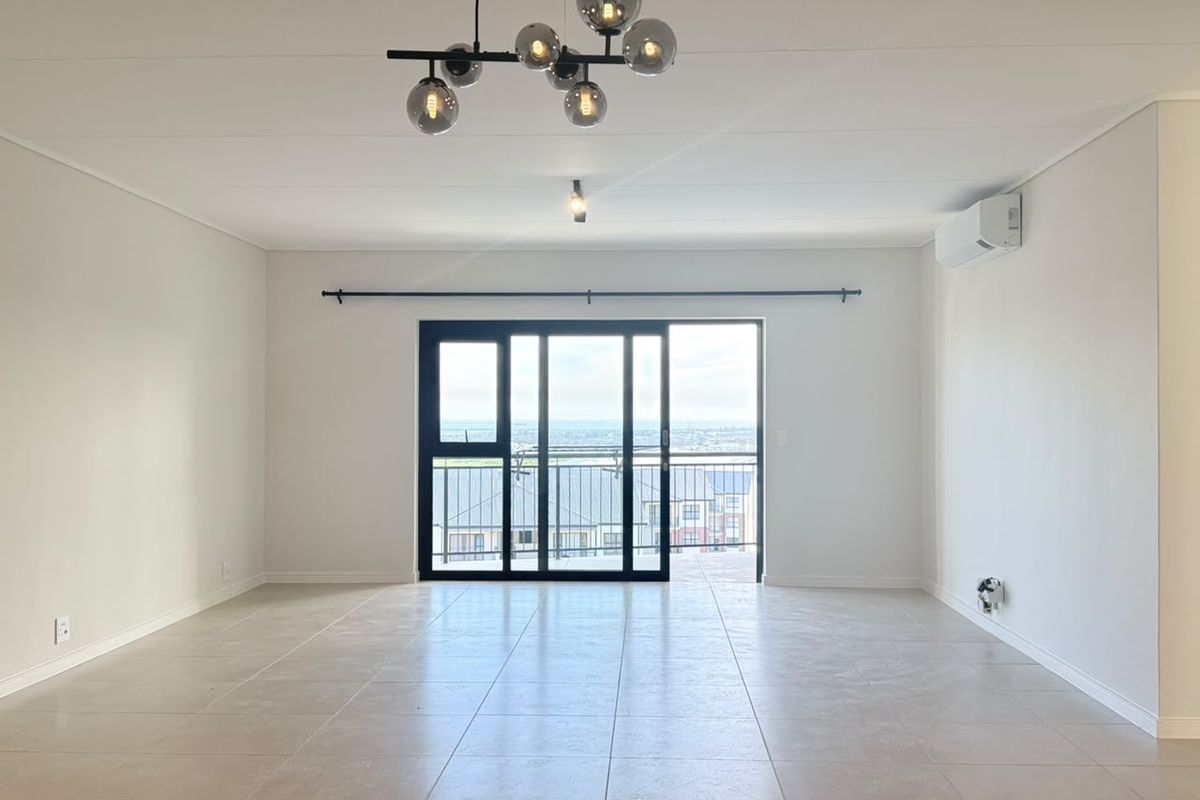 A bright, open-plan living area featuring neutral tiled flooring, a modern pendant light, and a sliding glass door leading to a balcony.