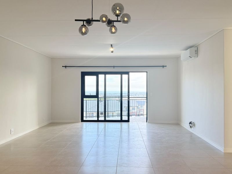 A bright, open-plan living area featuring neutral tiled flooring, a modern pendant light, and a sliding glass door leading to a balcony.