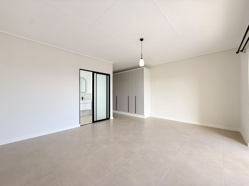 A bright, modern bedroom featuring neutral tiled flooring, built-in cupboards, and a sleek sliding door leading to an en-suite bathroom.