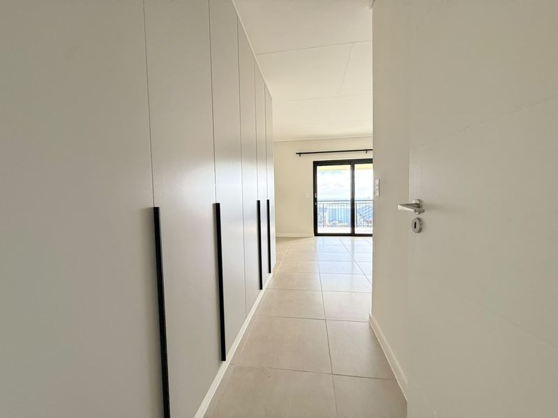 A bright, modern hallway featuring floor-to-ceiling built-in cupboards with sleek black handles, leading towards a sliding glass door with balcony access.