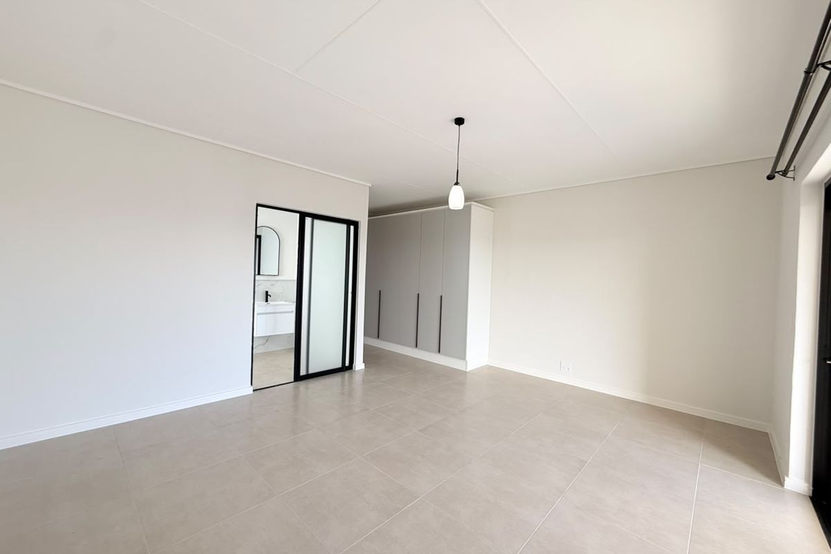 A bright, modern bedroom featuring neutral tiled flooring, built-in cupboards, and a sleek sliding door leading to an en-suite bathroom.