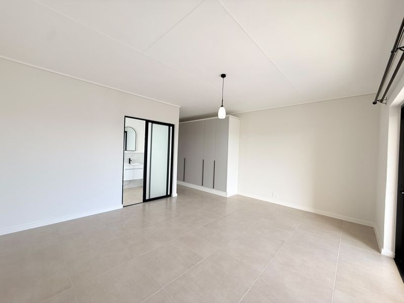 A bright, modern bedroom featuring neutral tiled flooring, built-in cupboards, and a sleek sliding door leading to an en-suite bathroom.