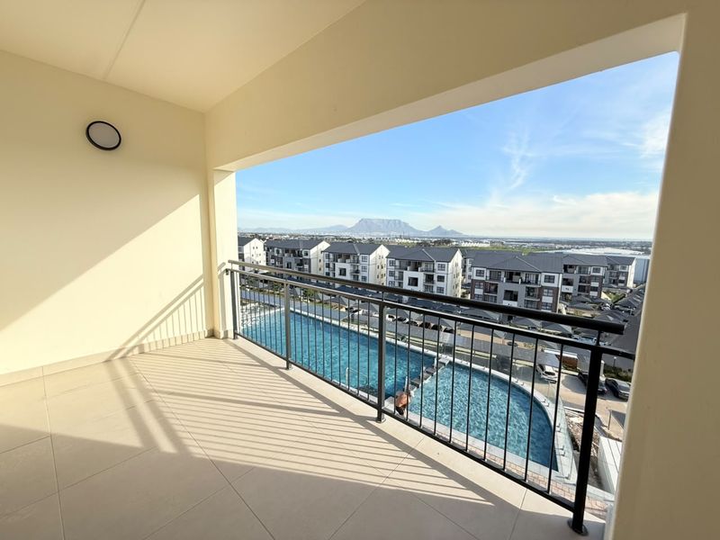 A spacious covered balcony featuring tiled flooring and a metal balustrade, offering elevated views over the complex swimming pool and surrounding landscape.