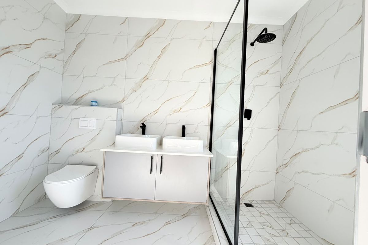 Modern bathroom featuring a double vanity, wall-mounted toilet, and a spacious walk-in shower with a black-framed glass screen and marble-look tiling.