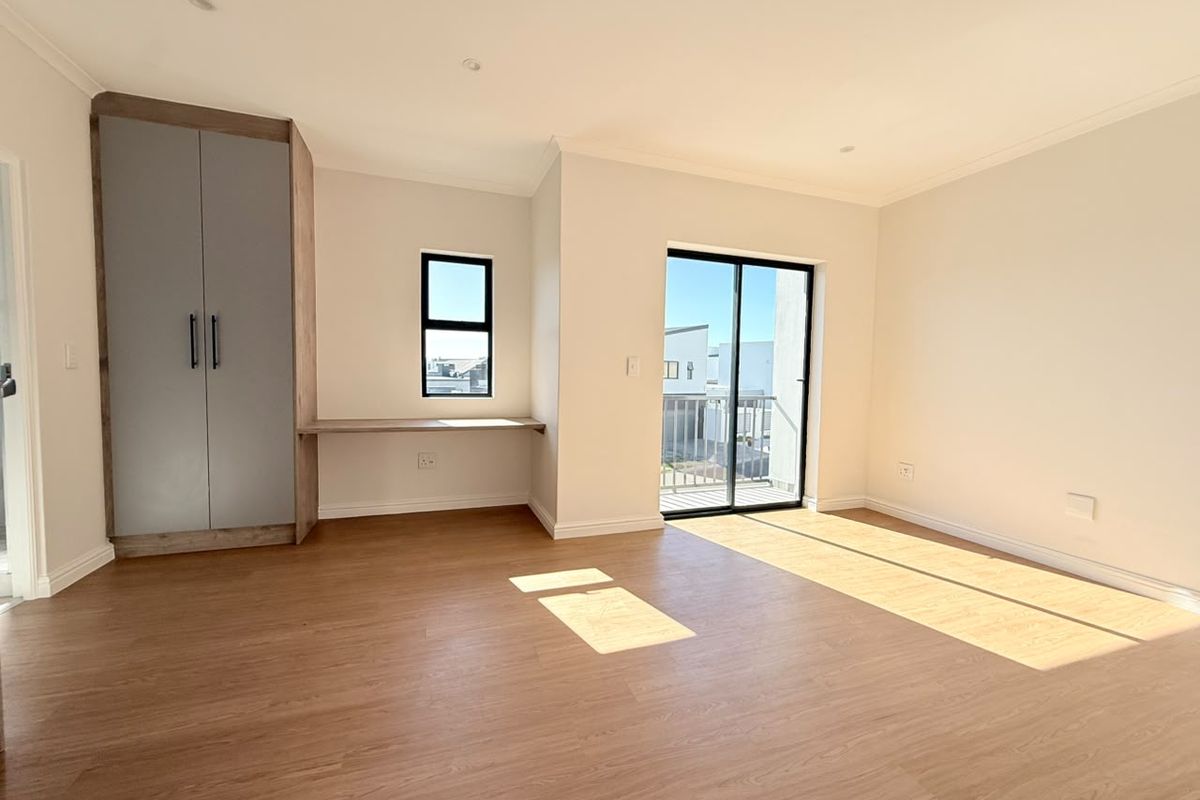 A bright, modern bedroom featuring laminate flooring, a built-in wardrobe with an integrated desk, and a sliding door leading to a balcony.