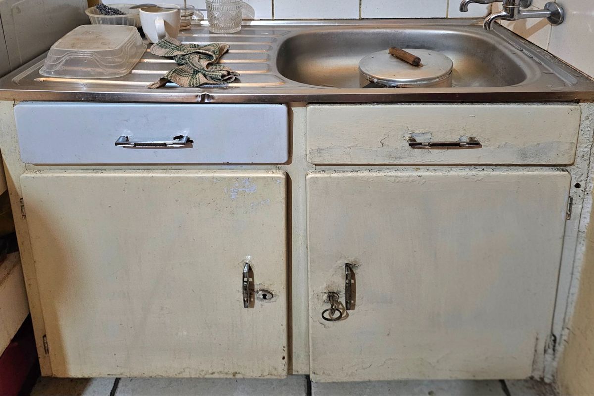 A dated kitchen sink area featuring a stainless steel basin and worn, painted cabinetry in need of refurbishment.