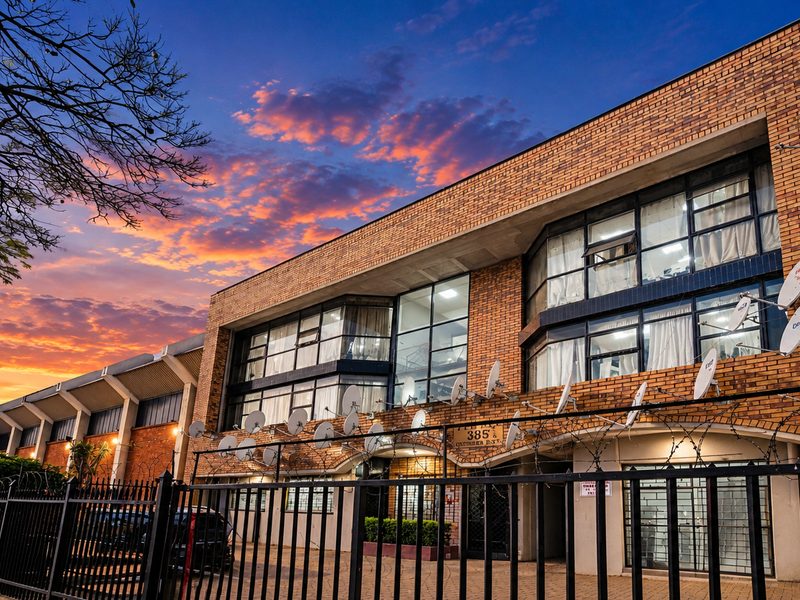 Front elevation of a multi-story commercial brick building featuring large windows, secure perimeter fencing with razor wire, and paved driveway access.