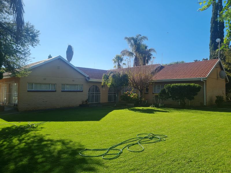 A front view of a single-story brick house with a tiled roof, featuring a large front lawn and security gates at the entrance.