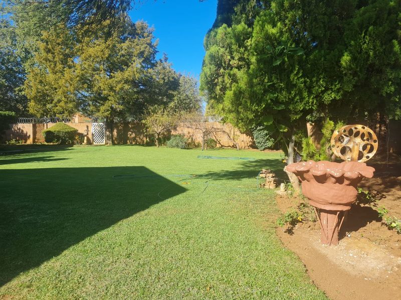 A well-maintained spacious garden featuring a manicured lawn, mature trees, and a secure brick boundary wall with security spikes.