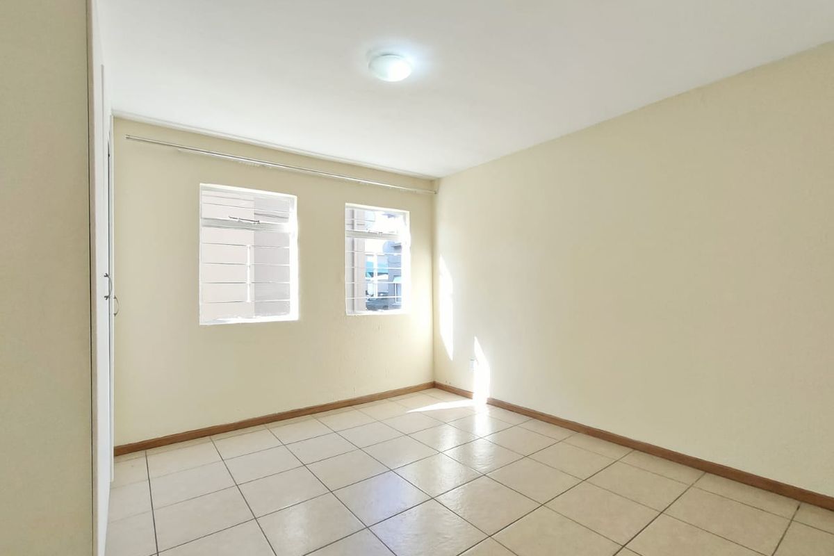A bright, empty bedroom featuring neutral tiled flooring, a built-in cupboard, and windows fitted with security bars.
