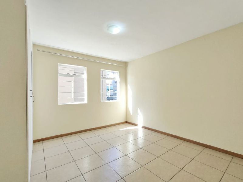 A bright, empty bedroom featuring neutral tiled flooring, a built-in cupboard, and windows fitted with security bars.