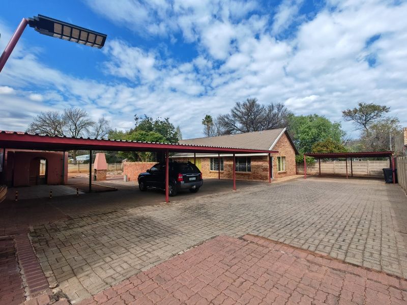 Spacious paved driveway featuring extensive covered carport parking, a brick-faced house, and a built-in outdoor fireplace area.