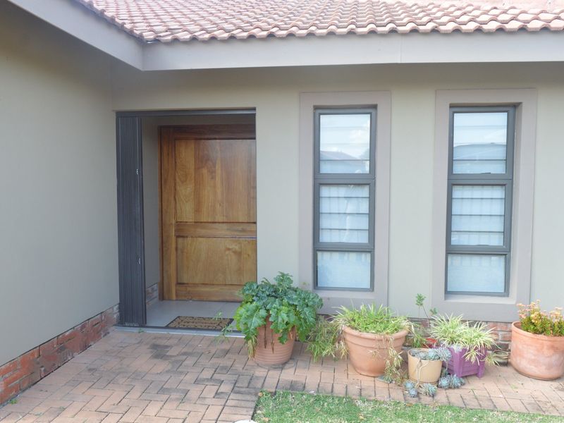 The front entrance features a solid wooden door, modern aluminium-framed windows, and a neat brick-paved patio area under a tiled roof.