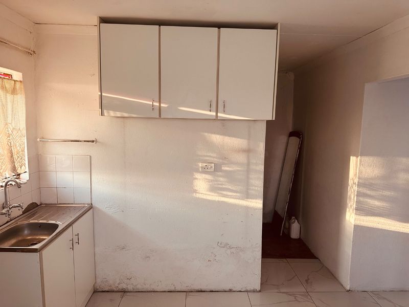 A compact kitchen area featuring basic built-in cabinetry, a stainless steel sink, and tiled flooring, requiring cosmetic updates to the wall surfaces.