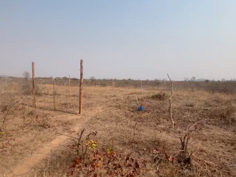 A view of a vacant residential plot featuring dry vegetation, wire perimeter fencing, and basic wooden fence posts.