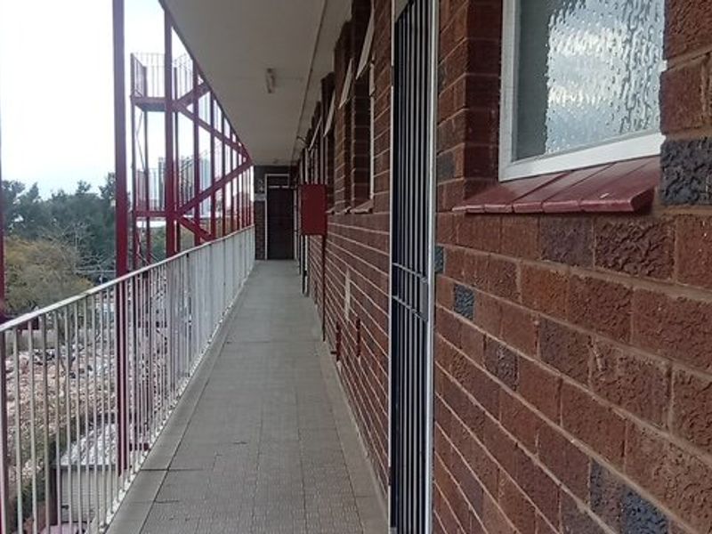 An exterior apartment building walkway featuring exposed brick walls, metal security gates, and a metal balustrade overlooking the surrounding area.