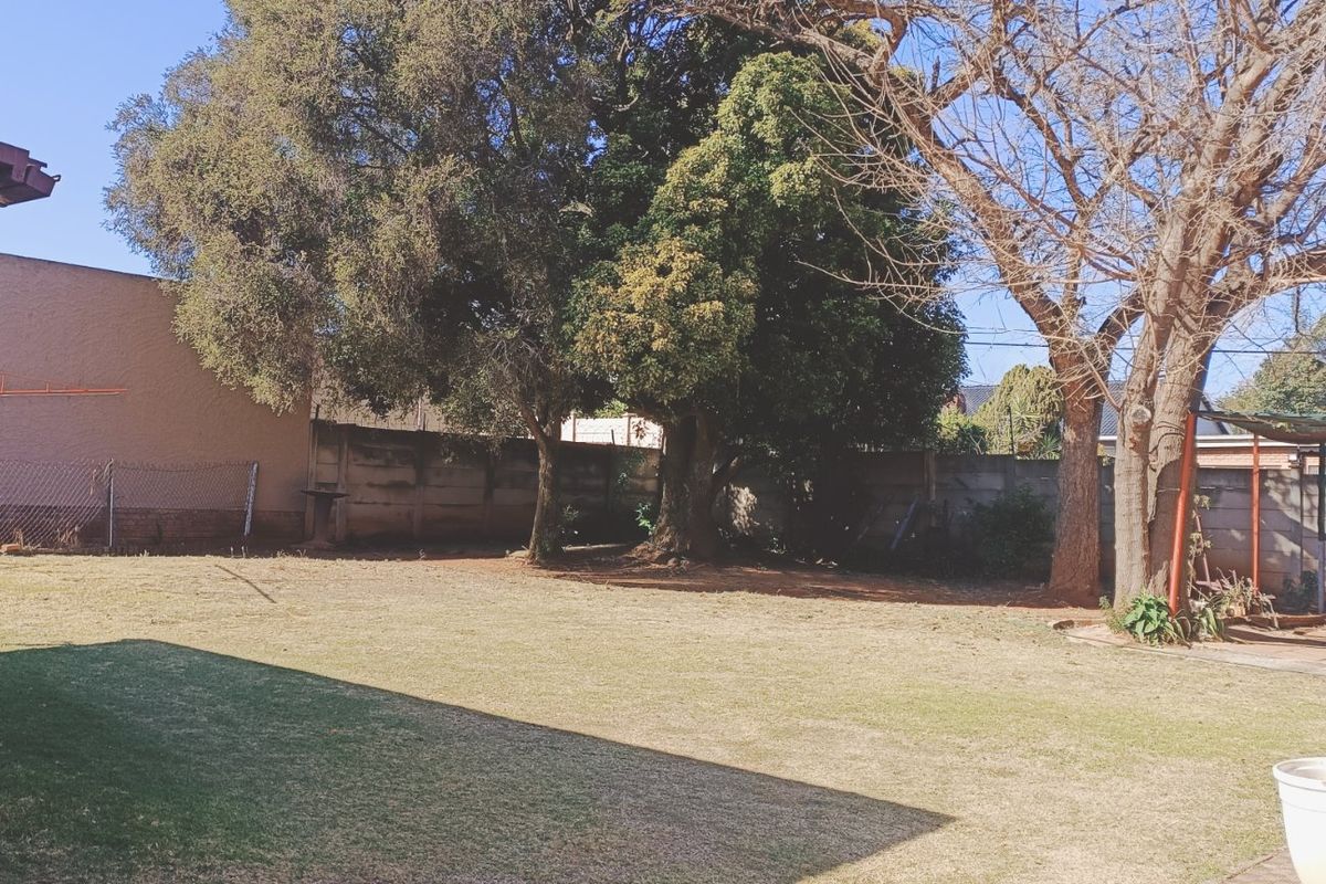 A spacious rear garden featuring a lawn, mature trees, and a precast concrete boundary wall with an adjacent covered patio area.