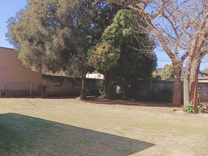 A spacious rear garden featuring a lawn, mature trees, and a precast concrete boundary wall with an adjacent covered patio area.