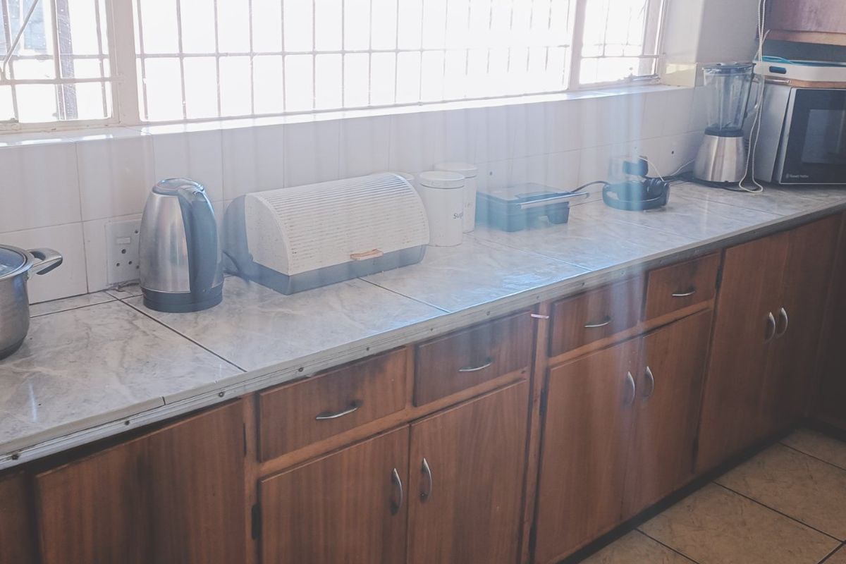 A functional kitchen featuring wooden cabinetry, tiled countertops, and a tiled backsplash, with security bars installed on the window.