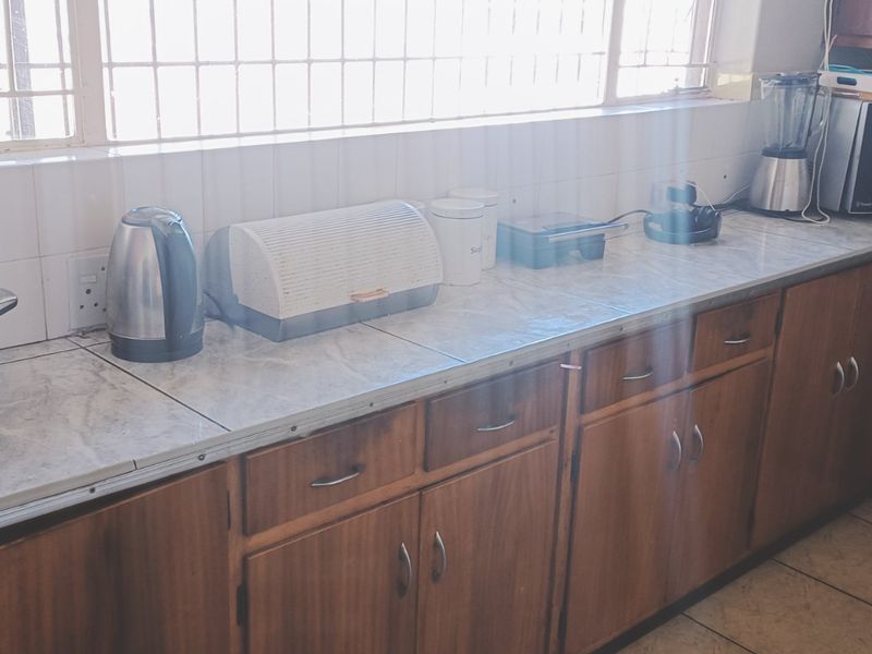 A functional kitchen featuring wooden cabinetry, tiled countertops, and a tiled backsplash, with security bars installed on the window.