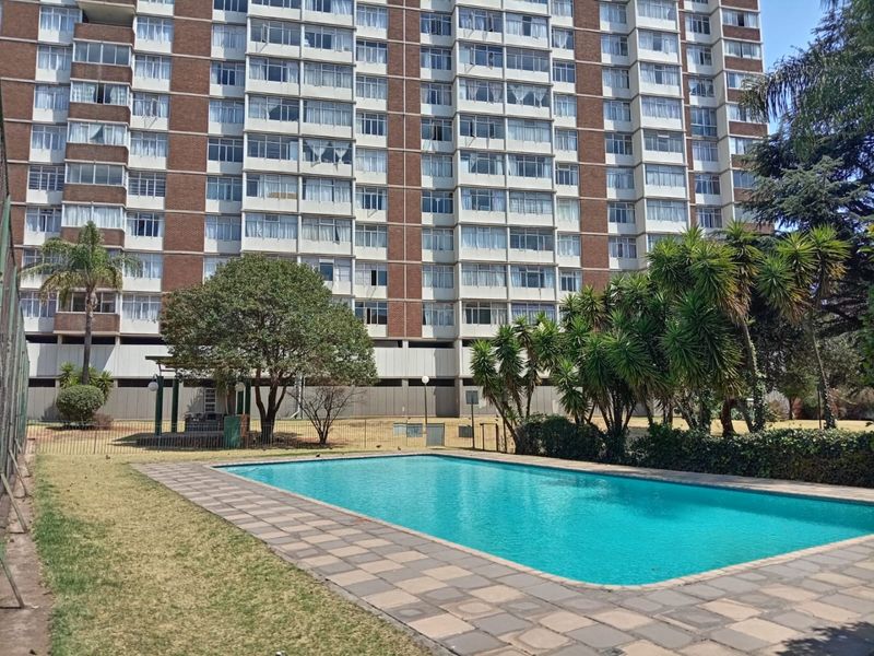 A communal swimming pool area set within a landscaped garden, featuring a paved surround and views of the apartment building facade.