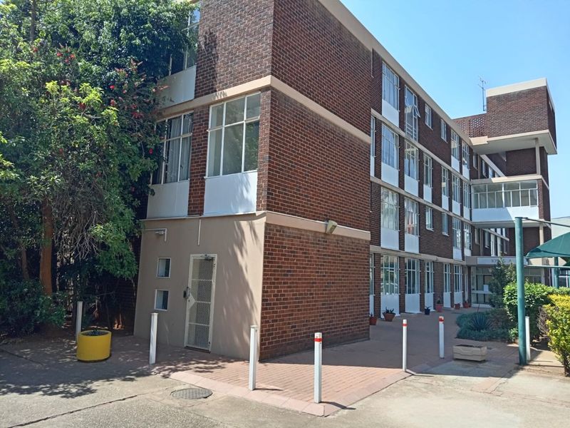 Exterior view of a multi-story brick apartment building featuring a paved entrance area, security gate, and established surrounding landscaping.