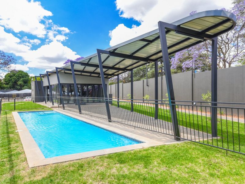 Well-maintained outdoor pool area featuring a modern metal pergola, paved walkway, and secure perimeter fencing within a landscaped garden setting.