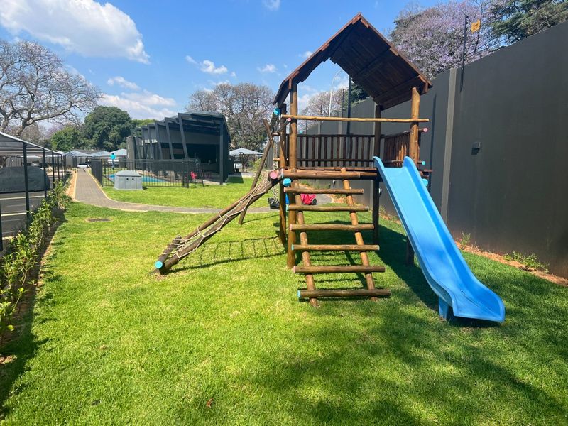A well-maintained communal garden area featuring a wooden playground structure with a slide, set against a boundary wall with paved pathways.